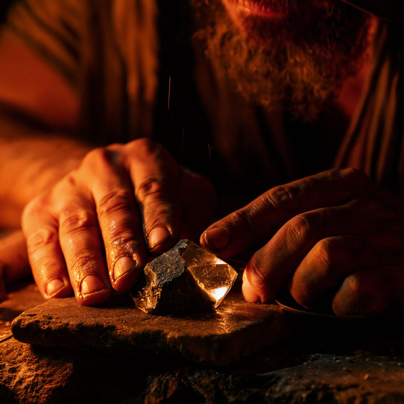 Hands working by candlelight, honing a rough dark crystal against a stone so that one face turns clear and faceted, fine sparks lifting into the dark.